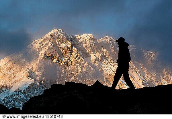 A man walks in silhouette on a ridge with Nuptse in the Himalayan background at sunset.