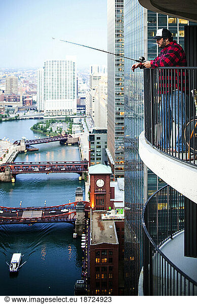 A man wearing a red plaid shirt pretends to fish from a balcony hundreds of feet above an urban river.