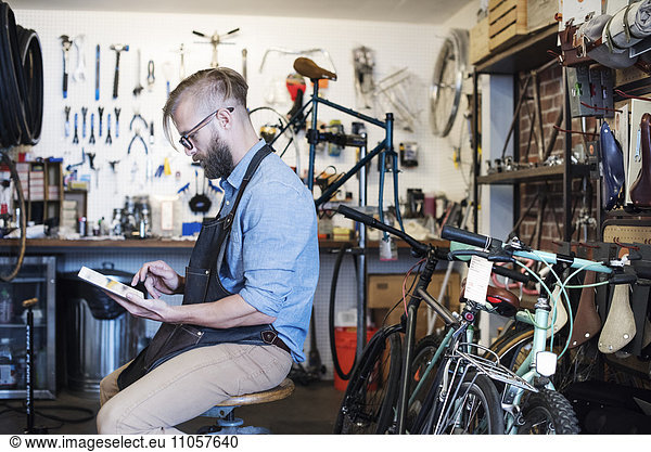 A man working in a bicycle repair shop  seated using a digital tablet.