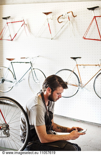 A man working in a bicycle repair shop sitting using his smart phone.