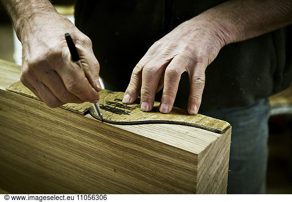 A man working in a furniture maker's workshop drawing around a shape on wood.