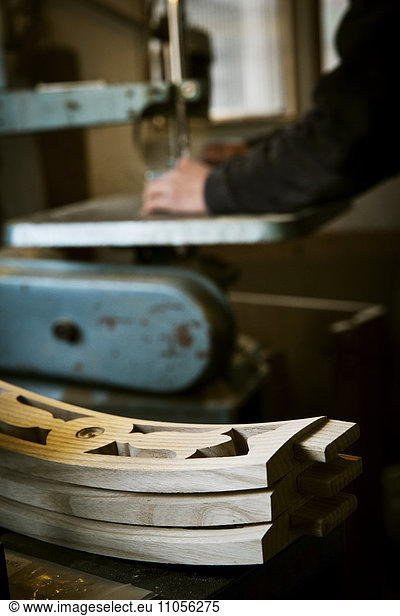 A man working in a furniture maker's workshop using a mechanical saw. Wooden chair backs stacked on a workbench.