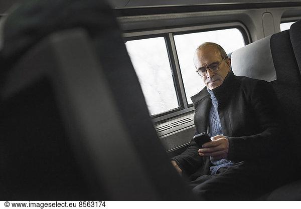 A mature man sitting by a window in a train carriage  using his mobile phone  keeping in touch on the move.