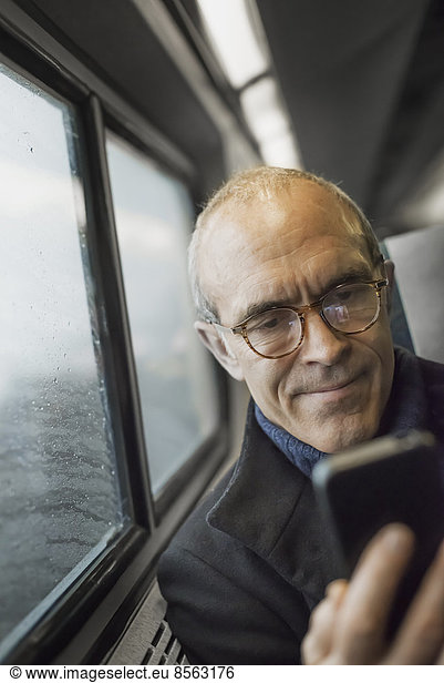 A mature man sitting by a window in a train carriage  using his mobile phone  keeping in touch on the move.