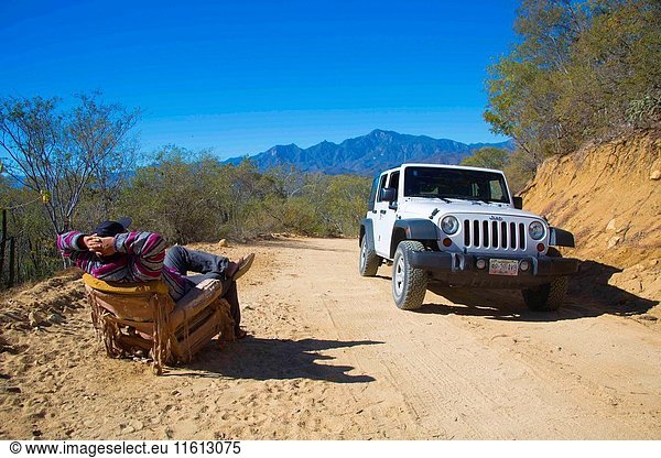 A mexican man is sitting on an old abandoned armchair  chair  with his arms behind his head  he is relaxing with the view. Front closeup of a white modern brand new Jeep with the savage  mountain  and dry land of Mexico behind it. We can see the mountains and imagine the off road fun the Jeep will have in this land.
