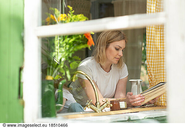 A mid woman looks at a recipe book by an open window in the kitc