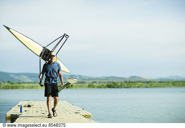 A middle-aged man carrying oars and a rowing shell on his shoulders  on a pontoon.