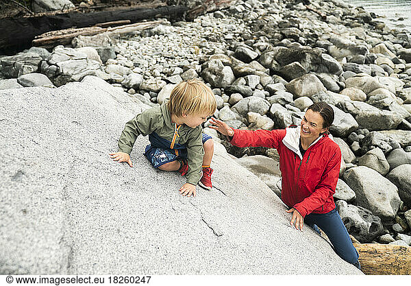 A mom helps her young son climb a boulder at a rocky beach