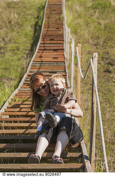 A mother and her young son sit together laughing on the lower steps of a long stairway leading to the top of a ski-jump at hilltop ski area Anchorage alaska united states of america