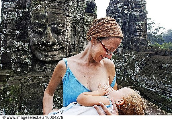 A mother breastfeeds her daughter baby in the Bayon temple. Angkor Thom. Cambodia. Angkor Thom was built as a square  the sides of which run exactly north to south and east to west. Standing in the exact center of the walled city  Bayon Temple represents the intersection of heaven and earth.Bayon is known for its huge stone faces of the bodhisattva Avalokiteshvara  with one facing outward and keeping watch at each compass point. The curious smiling image  thought by many to be a portrait of Jayavarman himself  has been dubbed by some the 'Mona Lisa of Southeast Asia. ' There are 51 smaller towers surrounding Bayon  each with four faces of its own.Bayon Temple is surrounded by two long walls bearing an extraordinary collection of bas-relief scenes of legendary and historical events. In all  there are are total of more than 11 000 carved figures over 1. 2km of wall. They were probably originally painted and gilded  but this has long since faded.