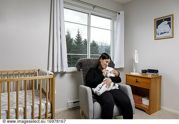 A mother sits in a rocking chair in a nursery and feeds her infant child with a bottle