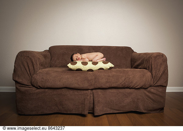 A naked newborn baby lying on his front  sleeping on a pillow on a brown couch.