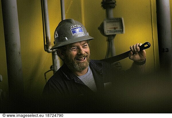 A oil drilling worker holds a large wrench over his shoulder and smiles.