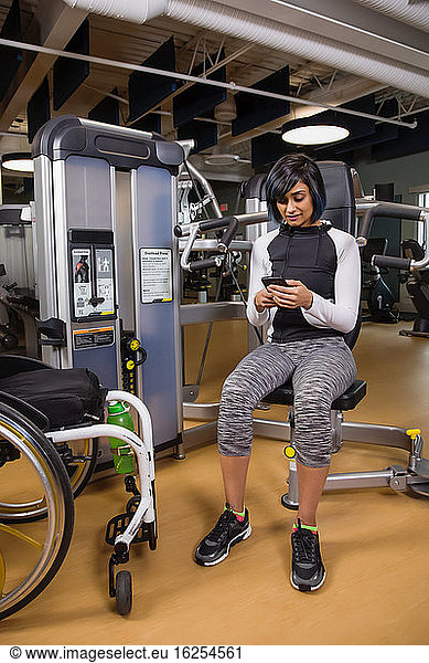 A paraplegic woman resting and texting on her smart phone after working out using an overhead press in a fitness facility; Sherwood Park  Alberta  Canada