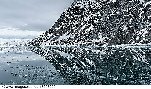 a partially snowy mountain is reflected in Arctic waters