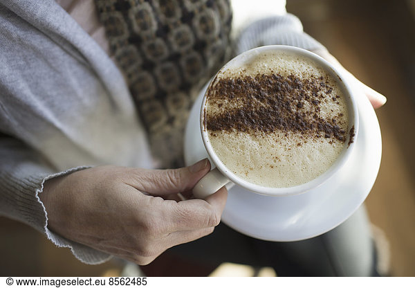 A person holding a full cup of frothy cappuccino coffee in a white china cup. Chocolate powder sprinkled in a pattern on the top. Coffee shop.
