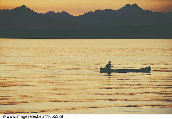 A person seated in a large Indian style canoe paddling across calm water at sunset.