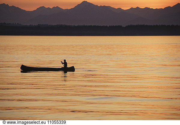A person seated in a large Indian style canoe paddling across calm water at sunset.