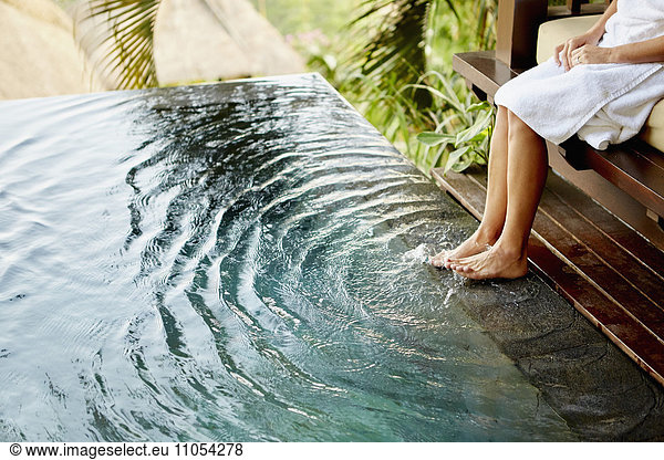 A person sitting on a bench with her feet in the shallow water of a pool  making ripples.