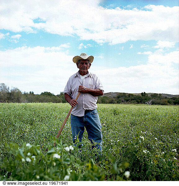 A portrait of a farmer standing in a field wearing a cowboy hat and holding a shovel.