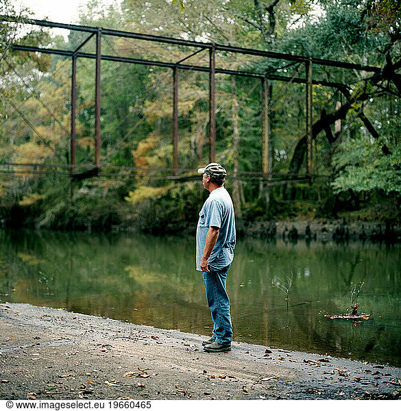 A portrait of a man next to a still river and a green forest.