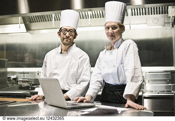 A portrait of a mixed race pair of chefs working with a laptop computer in a commercial kitchen.