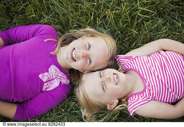 A Portrait Of Two Sisters Smiling. View From Above  Of Two Girls Lying On Their Backs On The Grass  Heads Together.