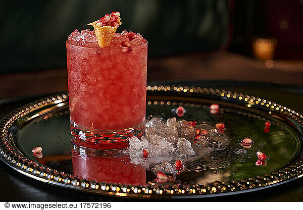 A red cocktail with pomegranate on a reflective tray.