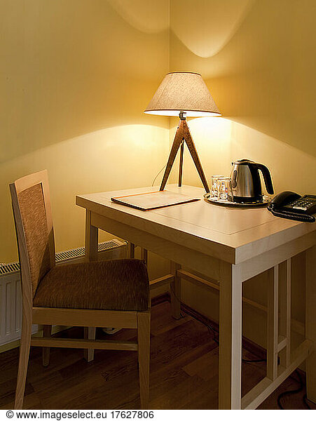 A room with a small desk and chair with a lit table lamp  tray and kettle and telephone.