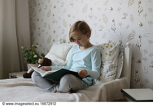 A Russian girl is reading a book on her bed in a bright room.