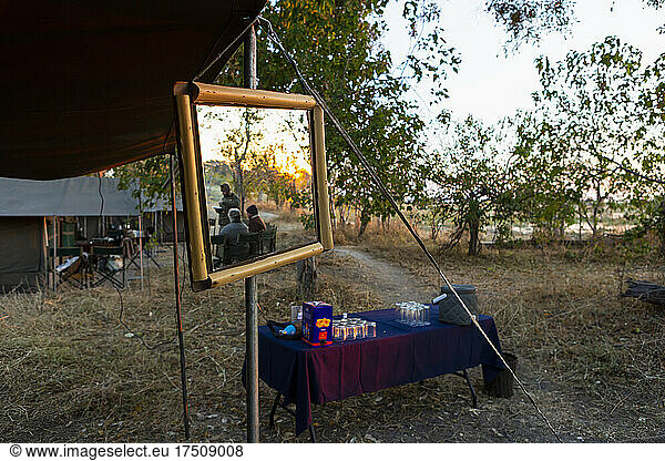 A safari camp  tents and drinks table  a large mirror on a tent pole  three people at sunset