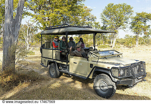 A safari jeep parked in the shade of a tree  people seated inside.
