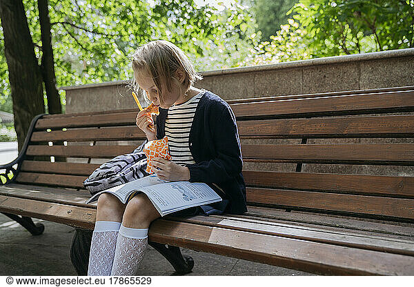 A schoolgirl eats a sandwich and does her homework in the park.