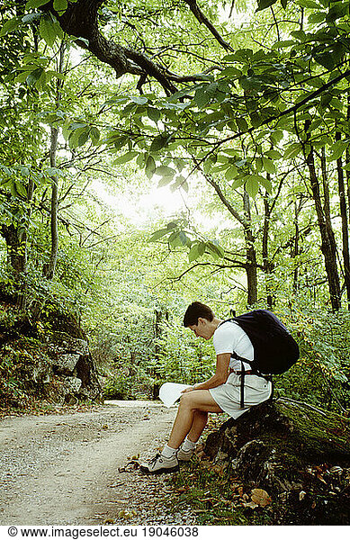 A seated female hiker reading a map along a wooded road in Tuscany  Italy.