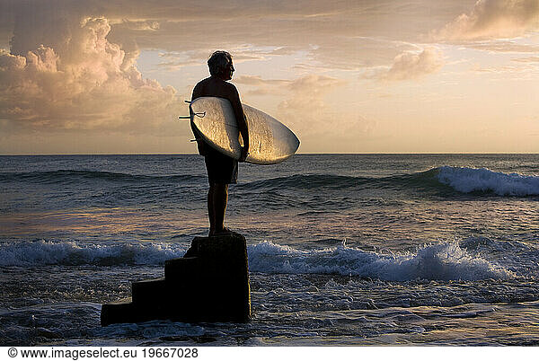 a senior surfer stands with his surfboard at sunset