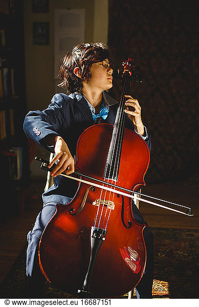 A serious boy in suit and bow tie with glasses plays cello at home