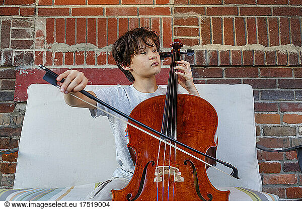 A serious boy plays cello outside against brick wall