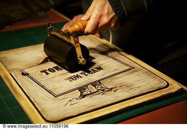 A sign maker rolling ink across the cut raised surface of linoleum preparing to print.