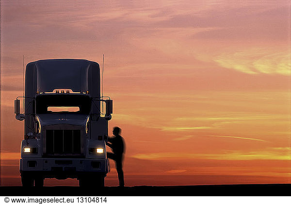 A silhouette of a truck driver getting into the cab of his commercial Class 8 truck tractor at sunrise.