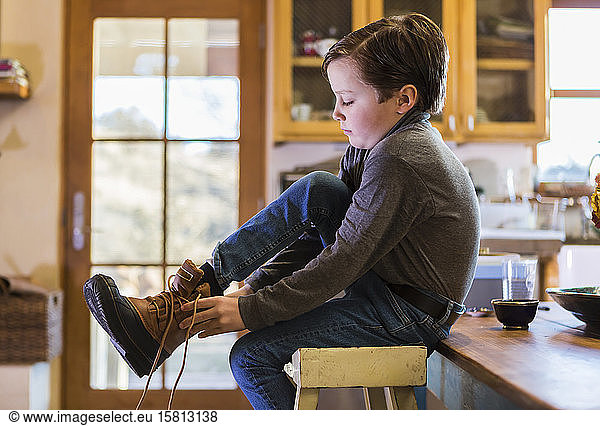 A six year old boy putting his boots on  sitting on a high stool.