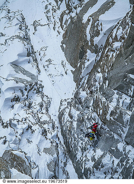 A skier and snowboarder hanging on a rock face while rappelling down to a couloir they plan on skiing.