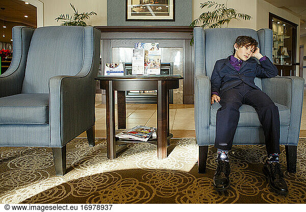 A small boy in a suit sits tiredly in a large chair in a hotel lobby