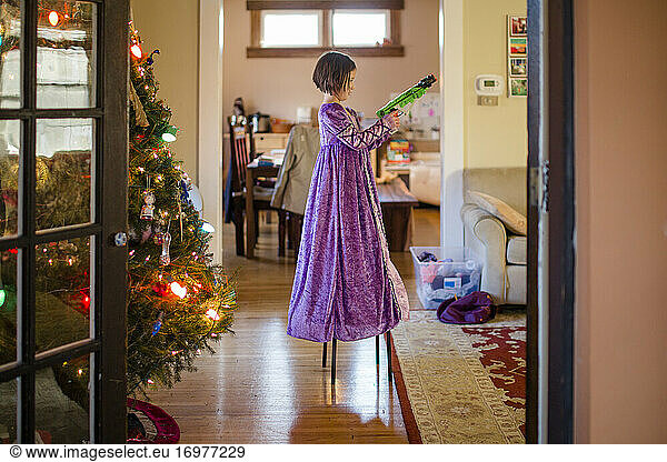 A small child in a long princess dress stands on chair with toy gun