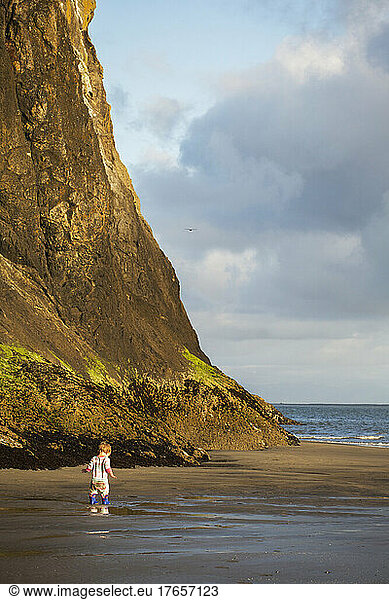 A small child in boots stands on a beach below a towering cliff