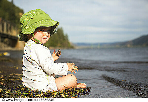 A small child in floppy hat sits in sand at water's edge