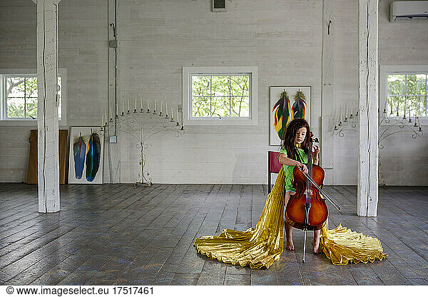 A small child in gold cape plays cello in large empty room