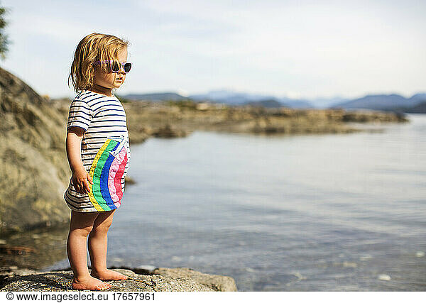 A small child in rainbow dress stands on rock at water's edge