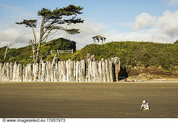 A small child sits on a wide beach with a driftwood sea wall