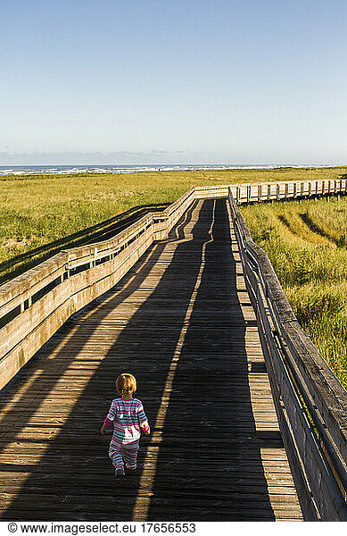 A small child walks down a long wooden boardwalk among grassy dunes