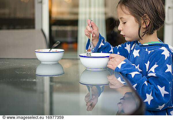 A small girl reflected in glass table eats breakfast outside